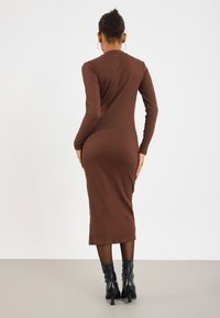 Donna che indossa un abito midi attillato lungo a maniche, colore marrone a coste, e stivaletti neri con tacco, in piedi con le spalle alla fotocamera.