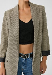 Beige rutmönstrad blazer med breda slag och svarta manschetter, kombinerad med en svart spetscroptop och högmidjade blåjeans som har ett svart bälte.