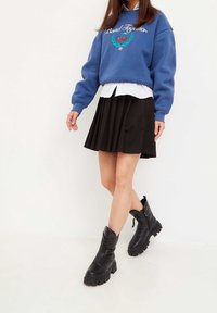 Sudadera azul con texto gráfico y diseño, camisa blanca con cuello debajo, falda negra plisada y botas negras hasta el tobillo con acabado texturizado.