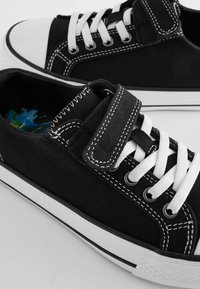 Sorte lærredssneakers med hvide snørebånd og gummitåkapper. Har en Velcro-rem og farverigt indvendigt foer. Kontrasterende hvid syning fremhæver designet.
