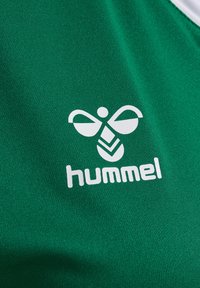 Logo de bourdon stylisé blanc et texte « hummel » sur tissu vert, probablement un vêtement de sport.