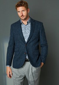Jūros jėgos spalvos herringbone tipo švarkas su dviem priekinėmis kišenėmis, rudais mygtukais ir įpjautais atvartais, dėvimas ant raštuoto šviesiai mėlyno marškinėlių ir pilkų klostuotų kelnių.
