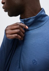 Άνδρας που ρυθμίζει το γιακά ενός μπλε μακρυμάνικου τοπ με υφή, με ένα μικρό κεντητό λογότυπο αλεπούς στο στήθος.