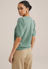 Pulover verde tricotat cu un model în zigzag, cu mâneci scurte pufoase și tiv ribbilt. Asortat cu pantaloni bej, largi.