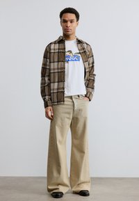 Νέος άνδρας που φοράει καφέ πουκάμισο με καρό σχέδιο πάνω από λευκό γραφικό μπλουζάκι, μπεζ παντελόνι φαρδιάς γραμμής και μαύρα παπούτσια, στέκεται μπροστά σε λείο τοίχο.