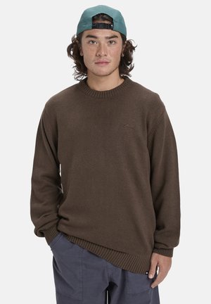 Pull en tricot marron avec un col rond à côtes et des manches longues, présentant un détail de logo subtil sur la poitrine. Tissu texturé.