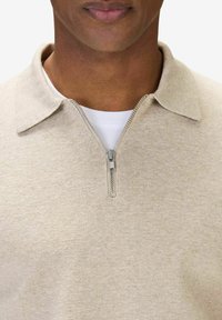 Beige zip-up polo shirt med en strukturerad tyg, med en klassisk krage och silver dragkedjedetalj. Lager ovanpå en vit t-shirt.