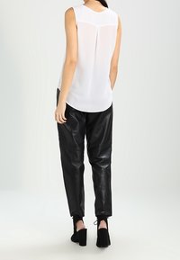 Witte mouwloze top met een plooi op de onderrug, gecombineerd met zwarte leren broek en zwarte hoge hakken, op een neutrale achtergrond.