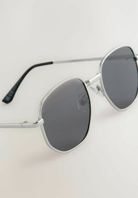 Gafas de sol de metal con un marco plateado, lentes hexagonales en gris oscuro y puntas de las patillas negras. Incluyen almohadillas nasales ajustables y un diseño elegante.