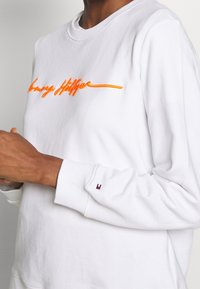 Person som bär en vit sweatshirt med orange kursiv text på bröstet och en liten marinblå och röd logotyp på vänster ärmslut.
