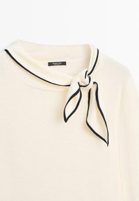 Pull crème à encolure ronde avec une écharpe bordée de noir nouée en un nœud au niveau du cou.