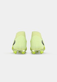 Par de botines de fútbol de color amarillo neón vistos desde atrás, mostrando cuellos altos de tobillo y logos de swoosh negros sobre un fondo blanco.