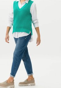 Tyrkysová pletená vesta s hlubokým výstřihem do V, nošená přes bílou košili; v kombinaci s krátkými modrými džíny a béžovými nazouvacími botami.