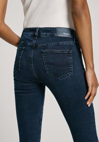 Jeans de mezclilla azul oscuro con una cintura de altura media. Las características incluyen dos bolsillos traseros y detalles de costura sutiles. Material suave, ajustado y elástico.