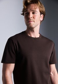 Brun crew-neck t-skjorte laget av glatt stoff, med korte ermer og et enkelt design, på en nøytral bakgrunn.