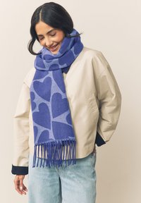 Sciarpa blu in misto lana con motivo a cuori, con bordi sfrangiati. Tessuto morbido e design caldo, drappeggiata su una giacca di colore neutro.