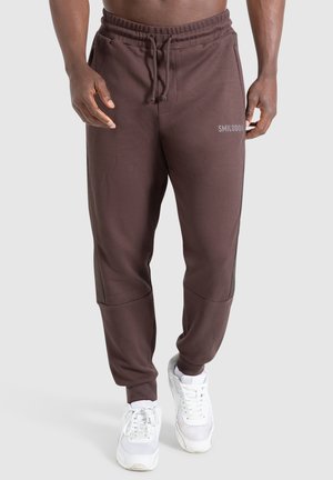 Brune sweatpants med en tekstureret overflade, elastisk talje, sidelommer og et sølvlogo på venstre lår. Parret med hvide sneakers.