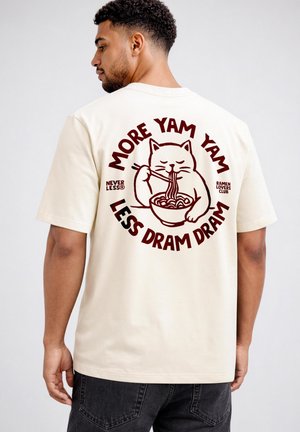 Man wearing cream shirt with graphic of cat eating ramen and text "More Yam Yam Less Dram Dram" on back.