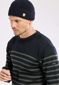 Homme portant un bonnet en tricot marine avec un patch d'ancre et un pull marine avec des rayures horizontales vertes et des boutons noirs sur l'épaule gauche.