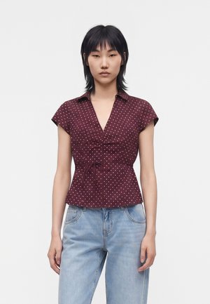 Frau trägt eine burgunderfarbene kurzärmelige Polka-Dot-Bluse mit vorderer Schleife und hellblaue Jeans, stehend vor weißem Hintergrund.