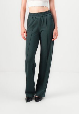 Femme portant un pantalon vert foncé taille haute à jambes larges avec coutures avant, associé à des chaussures noires pointues et un top court blanc.
