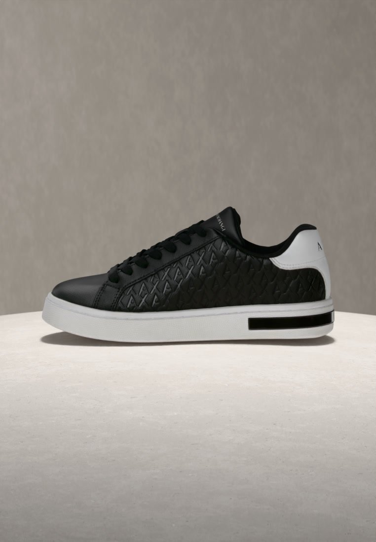 Baskets basses noires avec des panneaux latéraux texturés, un talon blanc, des lacets et une semelle blanche épaisse, sur un fond uni.
