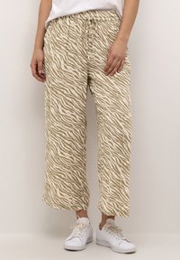 Personne portant un pantalon large à rayures zèbres beige et blanc et des baskets blanches, se tenant contre un fond clair.