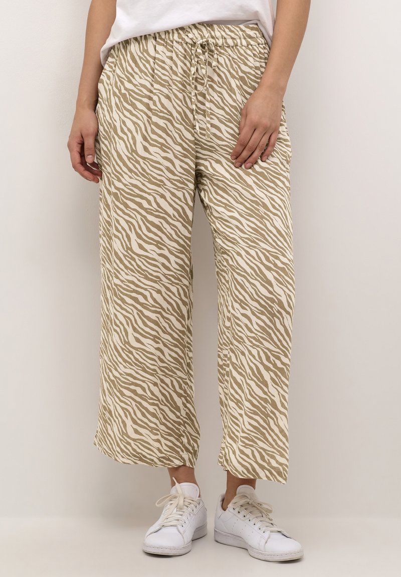 Personne portant un pantalon large à rayures zèbres beige et blanc et des baskets blanches, se tenant contre un fond clair.