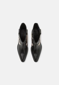 Botas de tobillo de cuero negro con punta afilada y diseño recortado en los laterales, que presentan acentos en color beige, tacón texturizado y detalles de costura.