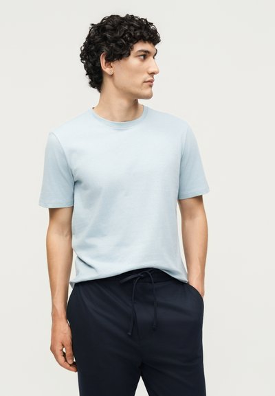 Άνδρας με καστανά σγουρά μαλλιά που φοράει ανοιχτό μπλε κοντομάνικο μπλούζα και σκούρο μπλε παντελόνι με κορδόνι, κοιτάζει δεξιά με τα χέρια στις τσέπες.