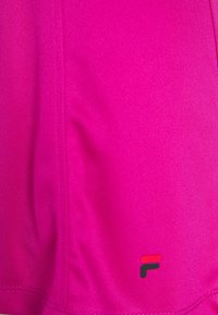 Top deportivo fucsia hecho de una tela suave y elástica con costuras visibles. Presenta un pequeño logotipo en rojo y negro en la parte inferior derecha.