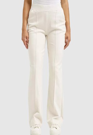 Frau trägt hoch taillierte, cremefarbene ausgestellte Hosen mit vorderen Nähten und weißen Sneakers vor einem schlichten grauen Hintergrund.