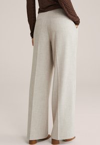 Naine, kes kannab beeže laiade säärega pükse ja pruuni pika varrukaga pealset, seisab ühe käega taskus ja on kaamerast eemale pööratud.