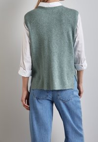 Ljusgrön ribbad stickad väst med rundad hals, öppna sidor och rak nederkant, lager på lager över en vit skjorta och blå denimjeans.