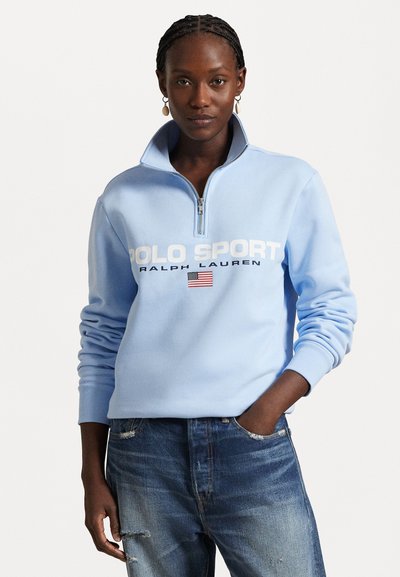 Jasnoniebieska bluza z suwakiem do połowy, wykonana z miękkiego materiału, z napisem "POLO SPORT" w białym kolorze oraz detalem amerykańskiej flagi. W połączeniu z postarzanym dżinsami.