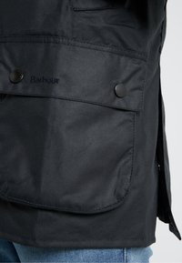 Zwarte waterdichte jas met grote zak, afgeronde zoom en knoopsluitingen. Heeft het geborduurde 'Barbour' logo op de zak.