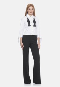 Camicia bianca con dettaglio a farfallino nero, bottoni dorati e maniche lunghe con polsini regolabili, abbinata a pantaloni neri aderenti.