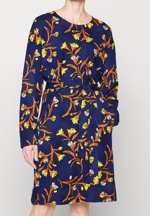 Kvinde iført en blå knælængde kjole med lange ærmer, bælte med bindebånd i taljen og et blomstermønster i orange og gul.