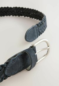 Vævet mørkeblåt ruskindsbælte med en sølvmetal spænde. Har en tekstureret overflade og syninger på remmens kanter.