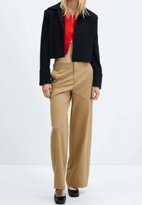 Zwarte cropped jas met knopen, gecombineerd met beige wijde broek. Rood geribbeld topje eronder. Glanzende zwarte flats maken de outfit compleet.