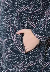 Modrá tkanina s bílými tečkami a růžovými větvičkami. Disponuje boční kapsou a má uvolněný střih, což zvýrazňuje hladkou strukturu textilu.