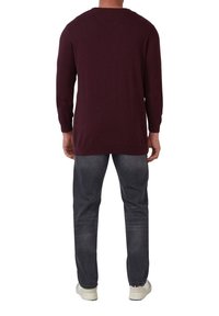 Bordeauxfarbener Langarm-Strickpullover mit gerippten Bündchen und Saum, kombiniert mit dunkelgrauen Jeans und weißen Sneakers. Glatte Textur im gesamten Pulli.