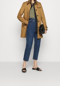 Beżowy trencz, oliwkowa bluzka, czarna koszula na guziki, niebieskie dżinsowe spodnie  cropped, czarne klapki oraz czarna torebka o fakturze.