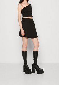 Top court noir à une bretelle, associé à une jupe noire évasée. Porté avec des bottes plateformes noires montant jusqu'au genou, avec une finition texturée.