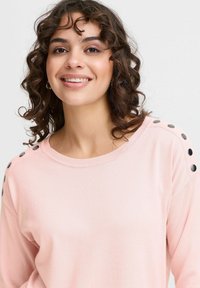 Pull rose clair en tissu doux, avec un grand col et des boutons noirs décoratifs sur les épaules. Manches longues, coupe décontractée.
