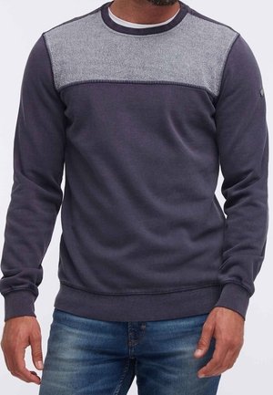 Muž nosící tmavě modrou mikinu se světle šedým pruhem přes ramena a světle modré džíny.