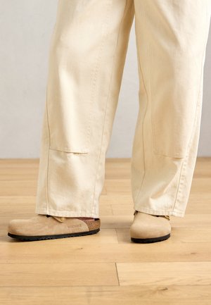 Beige wide-leg pants worn with tan suede Birkenstock clogs on a light wood floor.