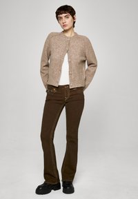 Jasnobrązowy dziergany kardigan z okrągłym dekoltem, zapinany na guziki, o fakturowanym wykończeniu, zestawiony z ciemnobrązowymi dzwonowatymi dżinsami i czarnymi butami.