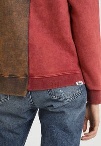 Bluza z kolorowymi blokami, z czerwonym górnym i brązowym dolnym fragmentem, z prążkowanymi mankietami i ściągaczem, zestawiona z niebieskimi jeansami.