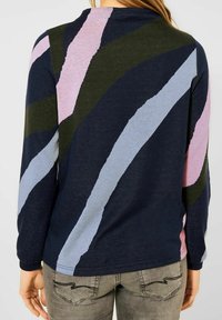 Osoba ubrana w navy niebieską bluzę z długim rękawem w szerokie ukośne paski w oliwkowym, jasnoniebieskim i różowym kolorze, do tego w szare dżinsy.